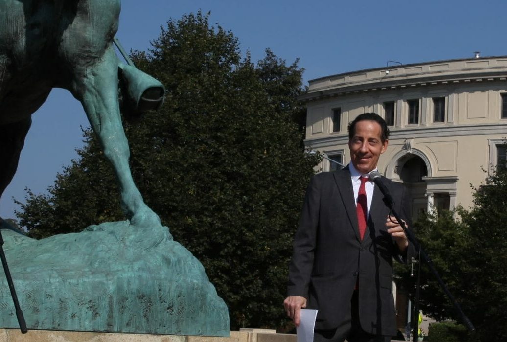Photo of Rep. Jamie Raskin (white man in black suit and red tie) standing next to iron monument of General Sheridan on a horse, speaking at a memorial event in 2019 with an embassy in the background.