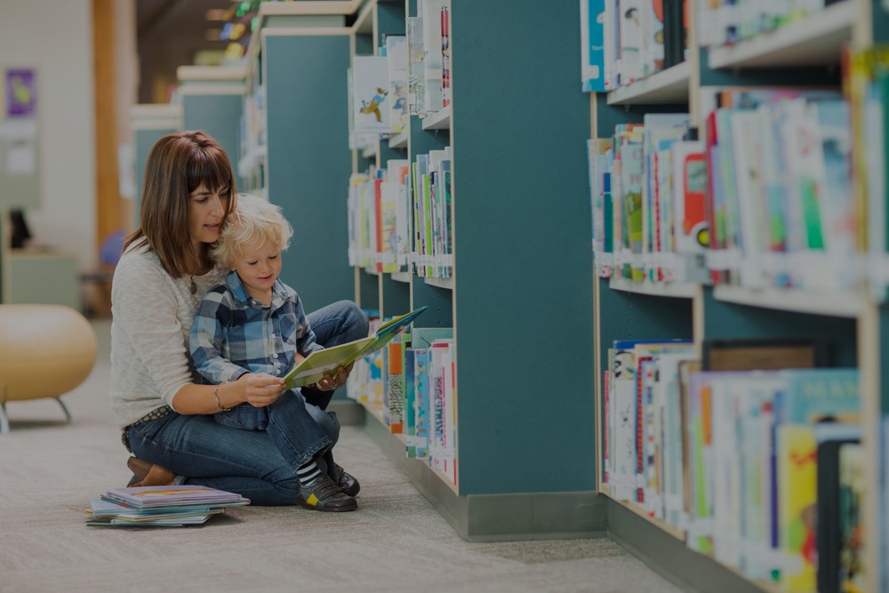 An image of a mother with her child in the aisle of a library. She is kneeling down with the child in front of her while she reads to the child.